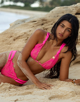 A woman with sun-kissed skin and wavy, tousled hair poses confidently on a beach, wearing a vibrant pink lace bikini from Beach Bunny Swimwear. The Lena Triangle Top features delicate lace overlay, adjustable knotted shoulder straps, and a gold-tone accent at the center. The matching Lena Tango Bottom has a cheeky cut with triple-strap side detailing, enhancing her curves. The backdrop showcases the rocks at the beach. Front view.